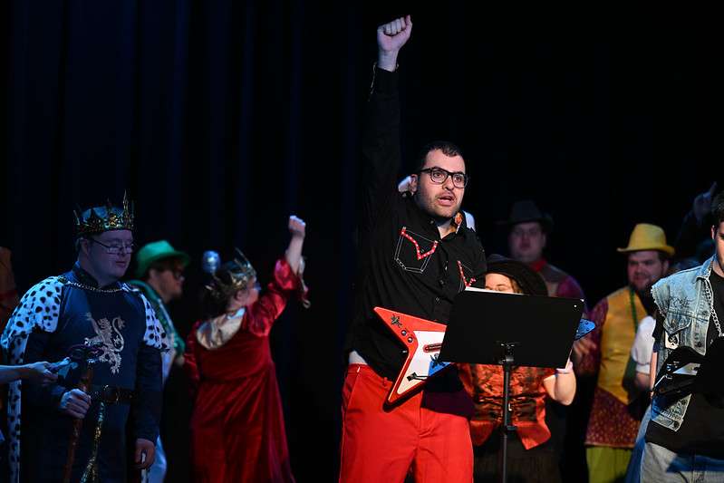 A performer stands center stage in a climactic moment, right fist raised high, wearing black and red stage clothing and glasses and holding a red electric guitar. The performer's expression is intense and fully present. A colorful ensemble fills the stage behind them in an array of character costumes — a royal blue robe and gold crown on the left, a flowing red dress, a yellow hat, a denim jacket, and others — suggesting a large-cast original production. Dark theatrical lighting frames the scene. A music stand is visible at the edge of the frame.